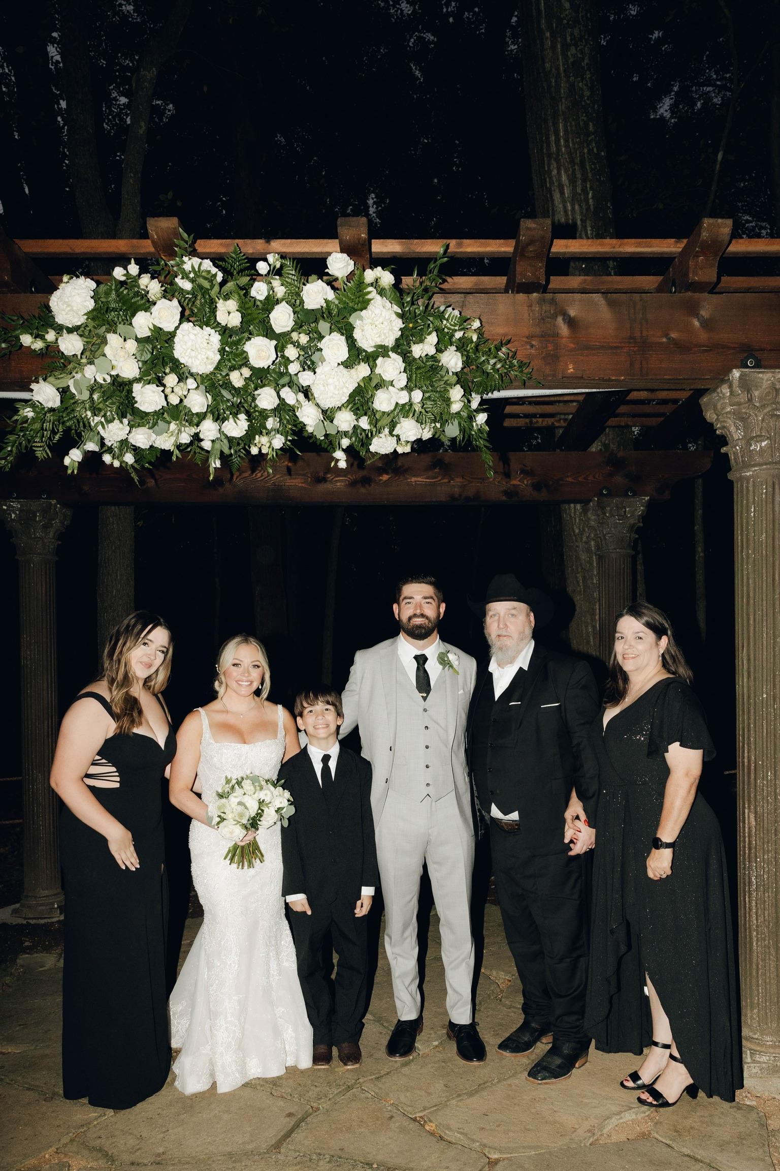 Guests and family pose together beneath a beautiful floral arrangement at a wedding.