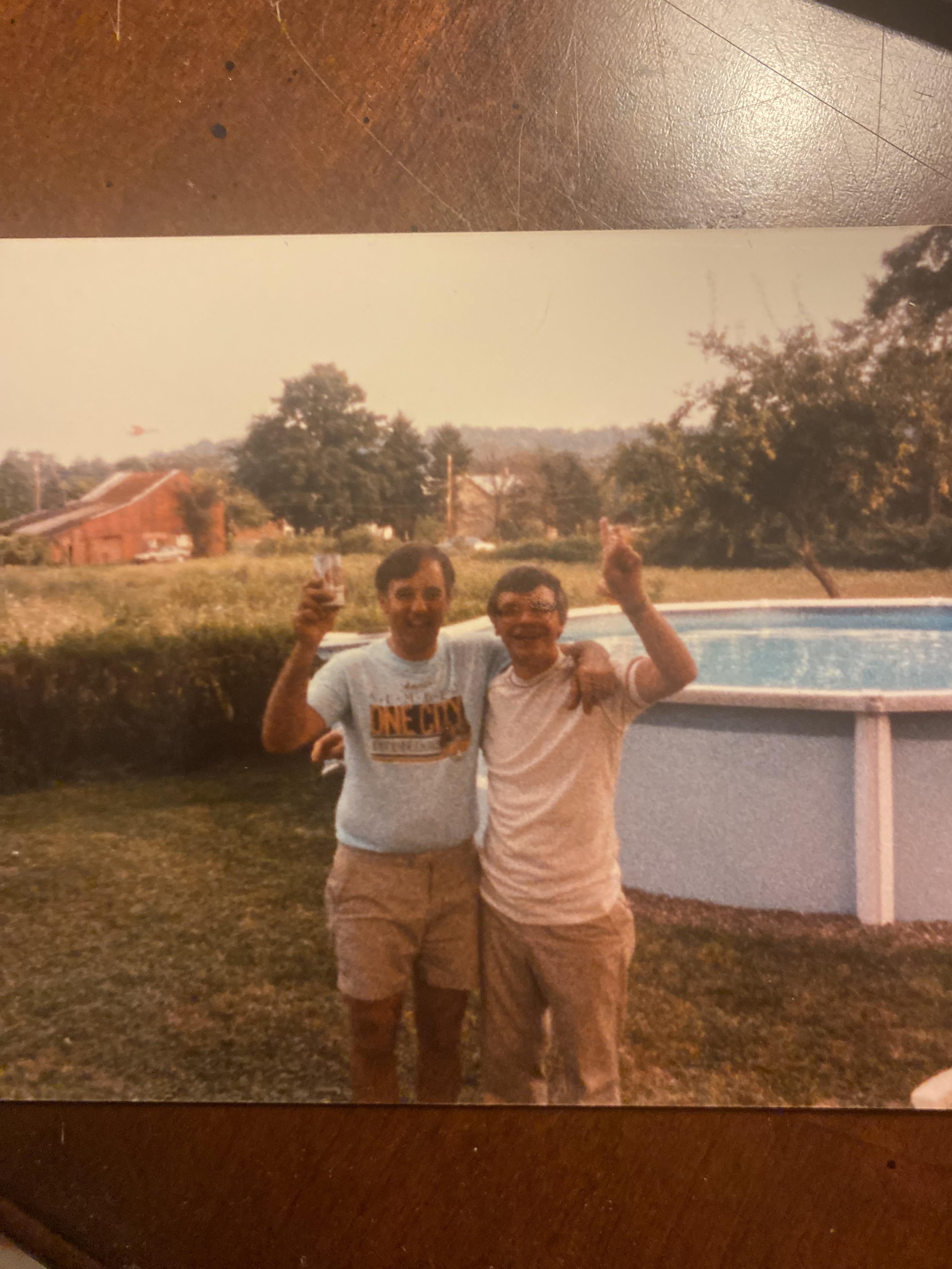 Two friends joyfully raise their glasses beside a pool in a grassy backyard under a clear sky.