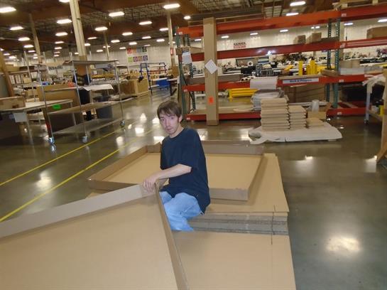 A person is sitting on the floor, assembling large cardboard pieces in a busy warehouse.
