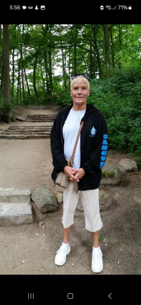 A woman in casual attire stands confidently on a wooded path surrounded by greenery.