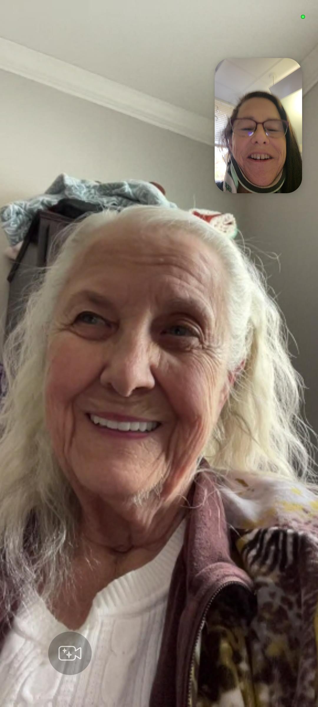 An elderly woman with long, white hair is smiling during a video call with a family member.