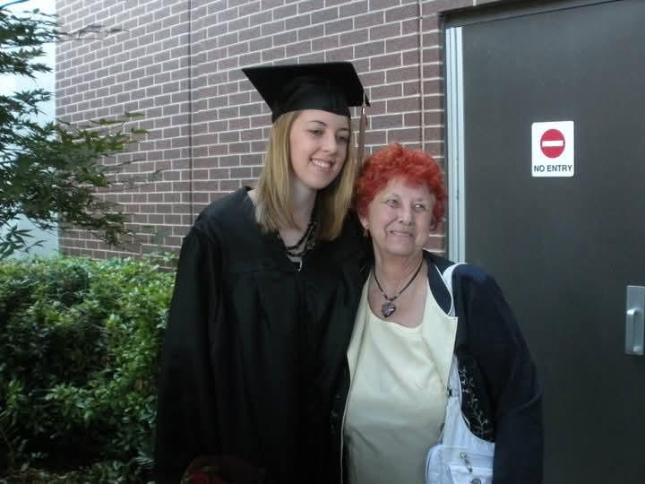 A young graduate poses proudly with a family member outside a brick building.