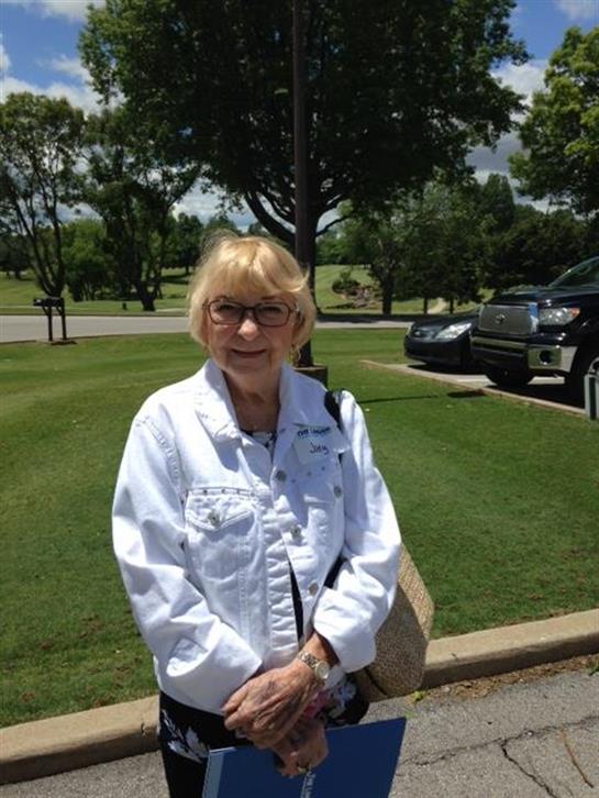 Elderly lady stands cheerfully in a park, wearing glasses and holding a binder, enjoying the day.