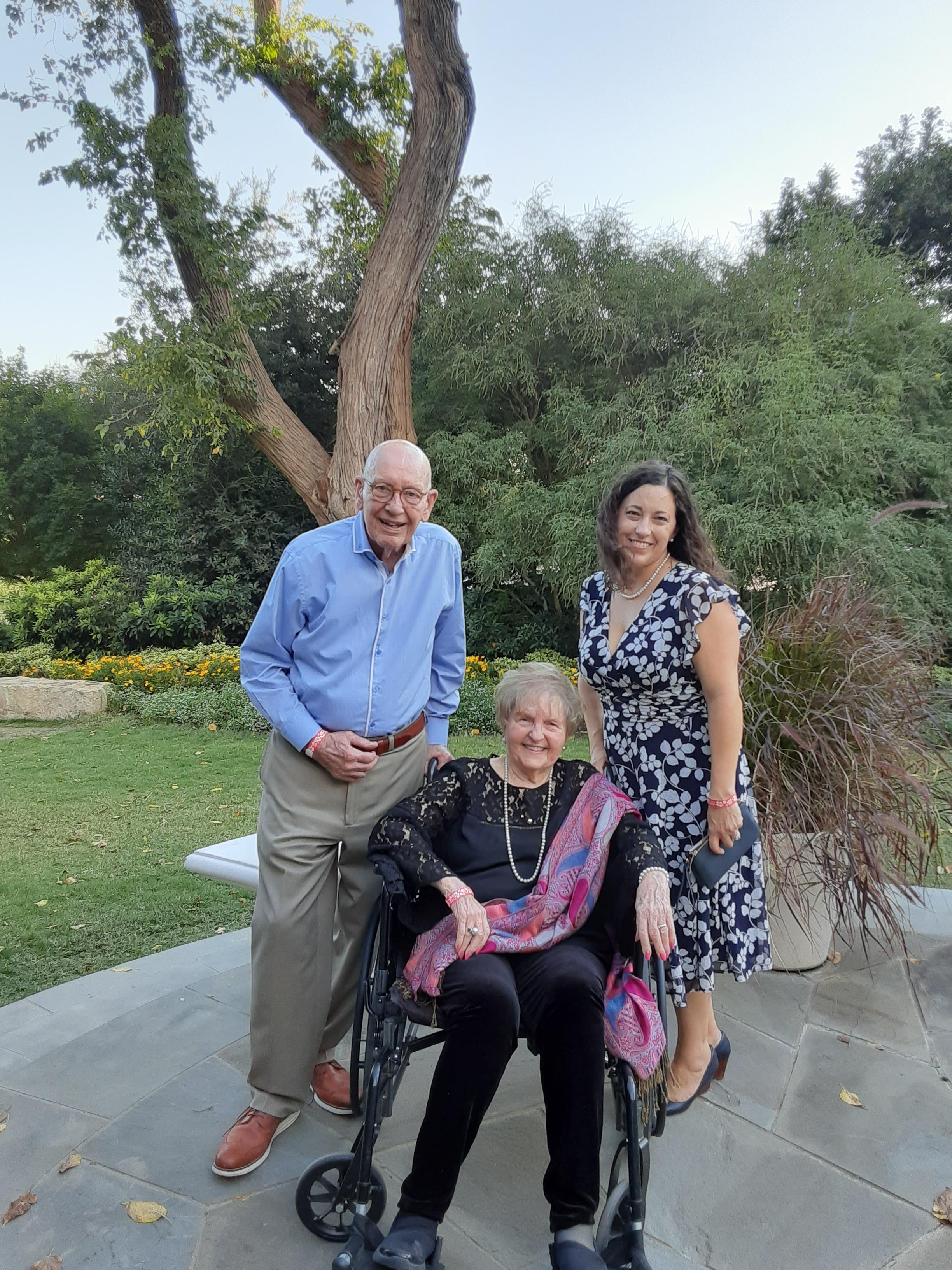 Three people are in a garden: one woman in a wheelchair and two adults standing nearby.