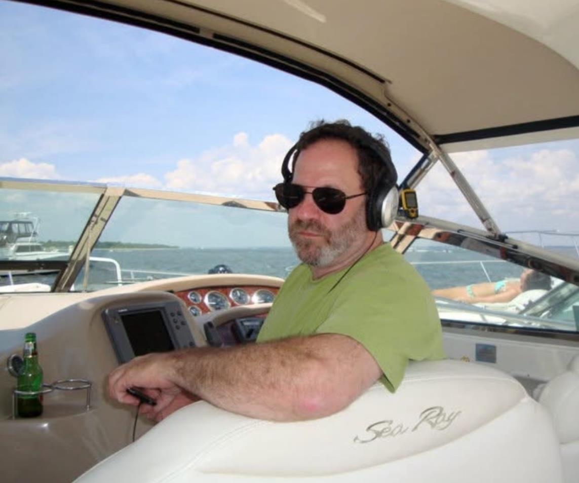 A man relaxes on a yacht, wearing headphones and sunglasses while steering the boat.