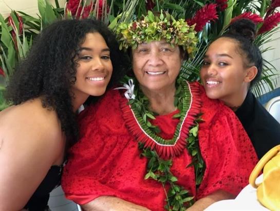 Elderly woman joins two smiling young women in traditional attire and floral crowns.