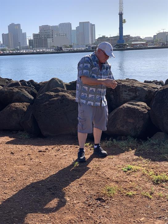 A man is standing on the shore, gazing at his phone with a city view behind him.