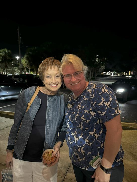 Two friends smile widely while standing outside a restaurant at night with cookies.