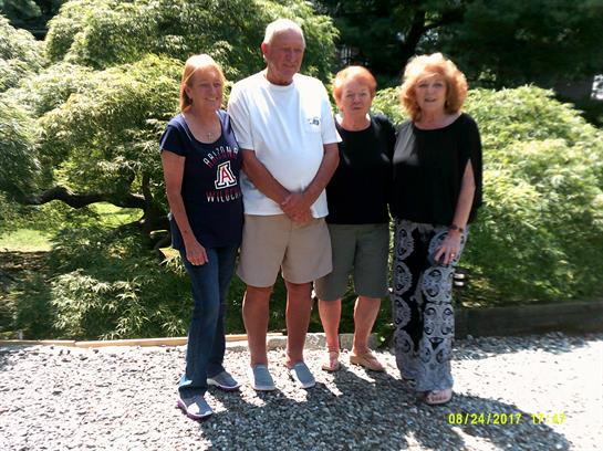 Four adults pose together in a lush green garden under bright sunlight, smiling happily.