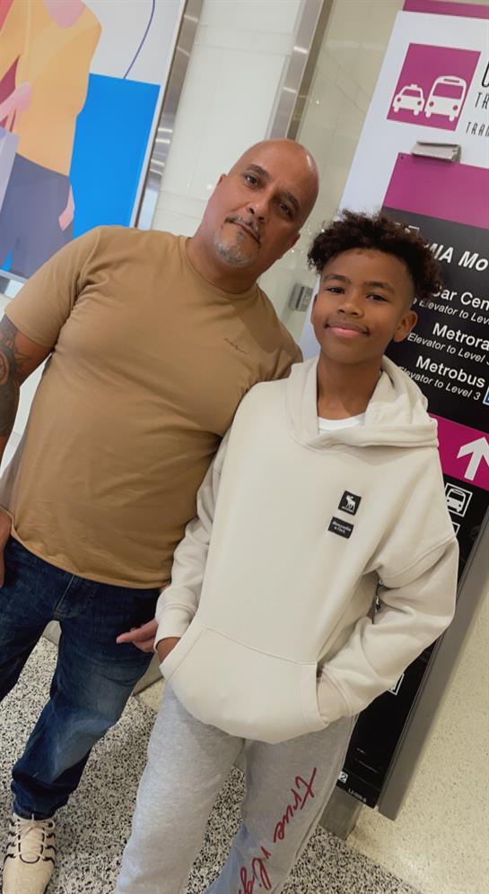 A man and a young boy smile while standing at a busy transportation location, surrounded by signage.