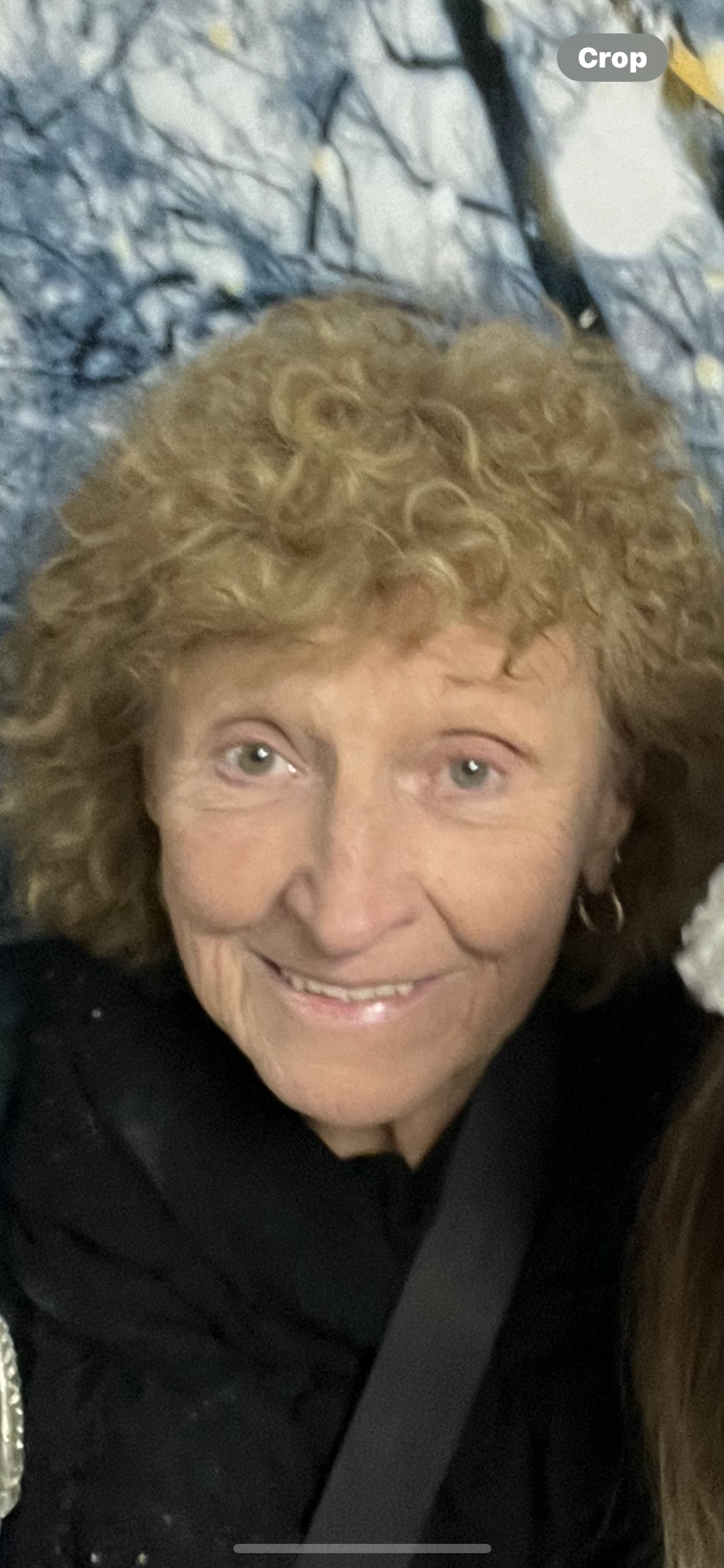 A cheerful elderly woman with curly hair smiles warmly in a cozy indoor environment.