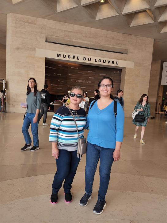 Two women stand happily inside the Louvre Museum, surrounded by fellow visitors.