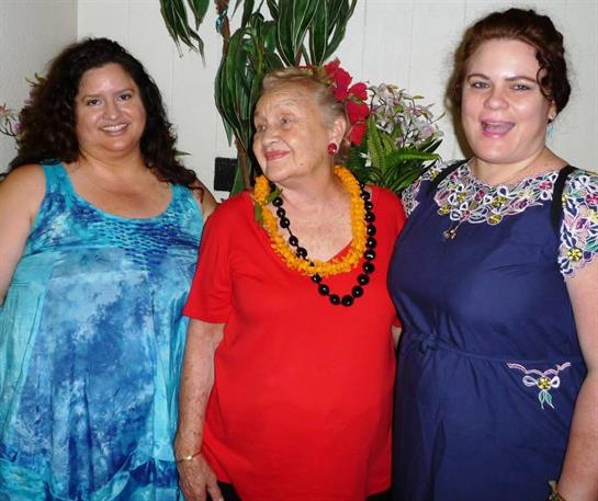 Three women smile as they gather in a colorful space filled with tropical plants and flowers.