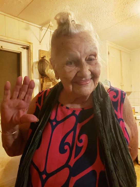 An elderly woman with a joyful expression waves from a cozy kitchen, surrounded by warmth.