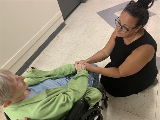 A caregiver kneels beside an elderly person in a wheelchair, holding hands and providing comfort.