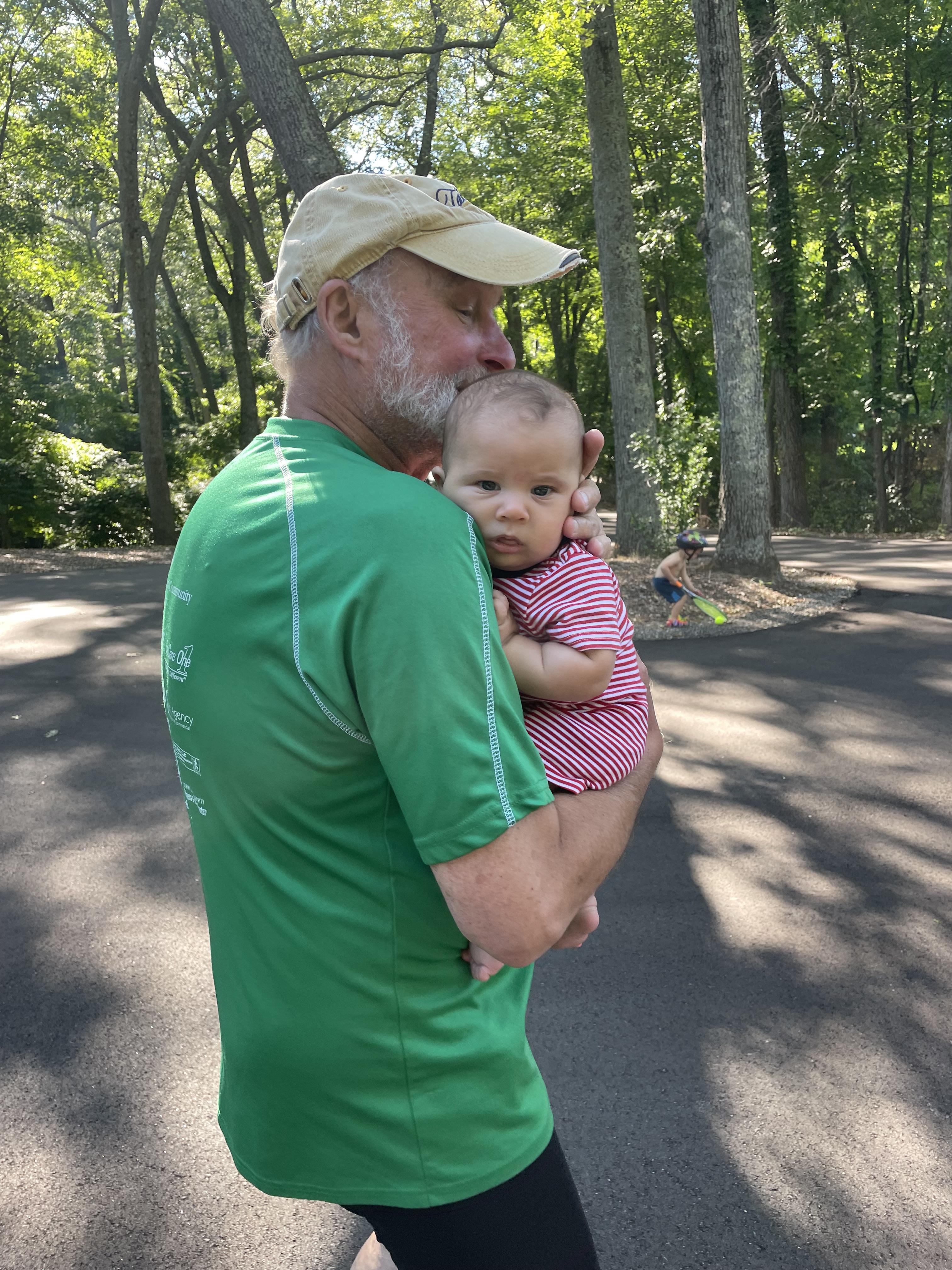A man holds a baby girl in his arms while walking through a park on a sunny day.