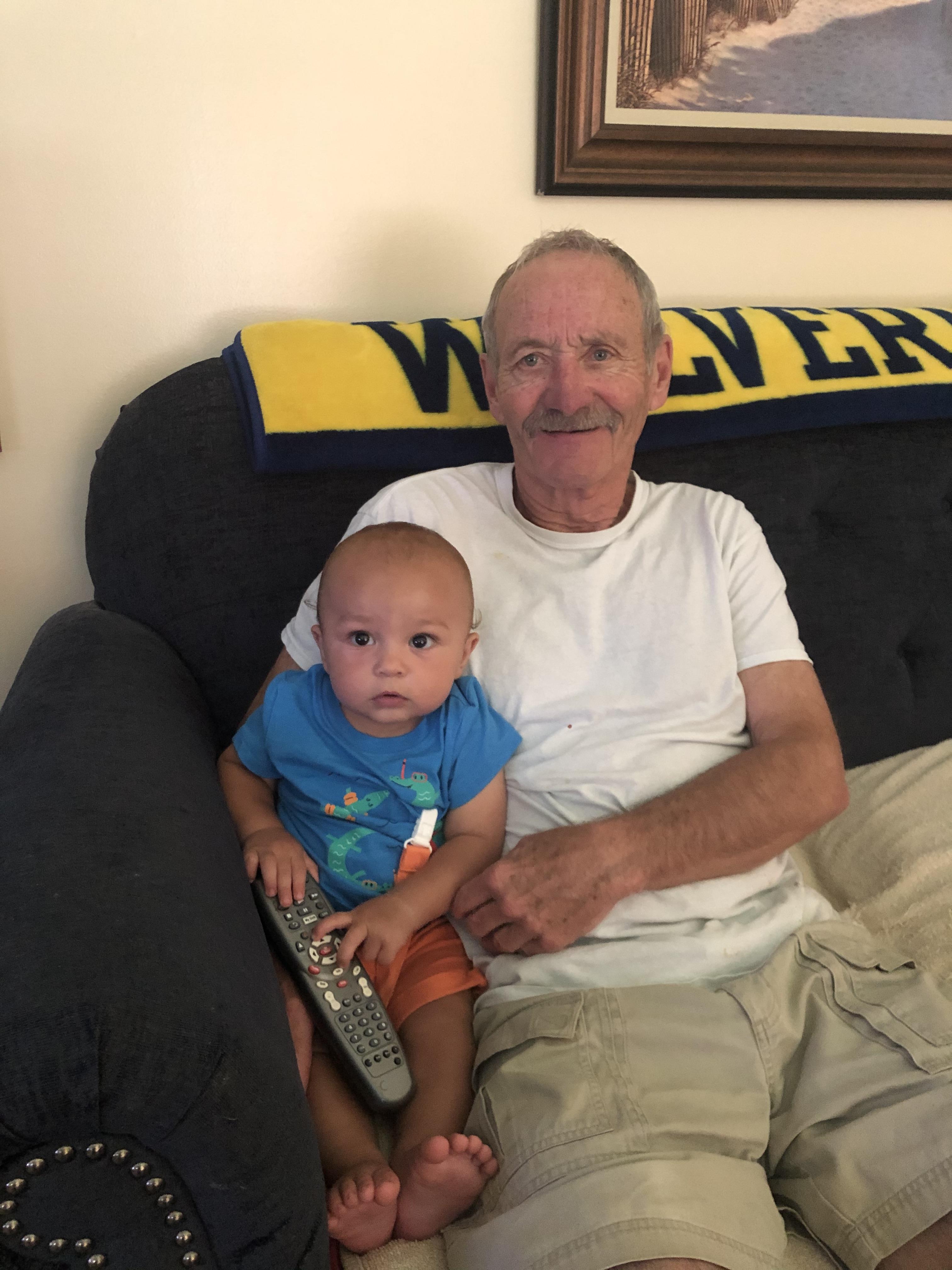 An elderly man and a baby sit together on a couch, sharing a moment of joy at home.