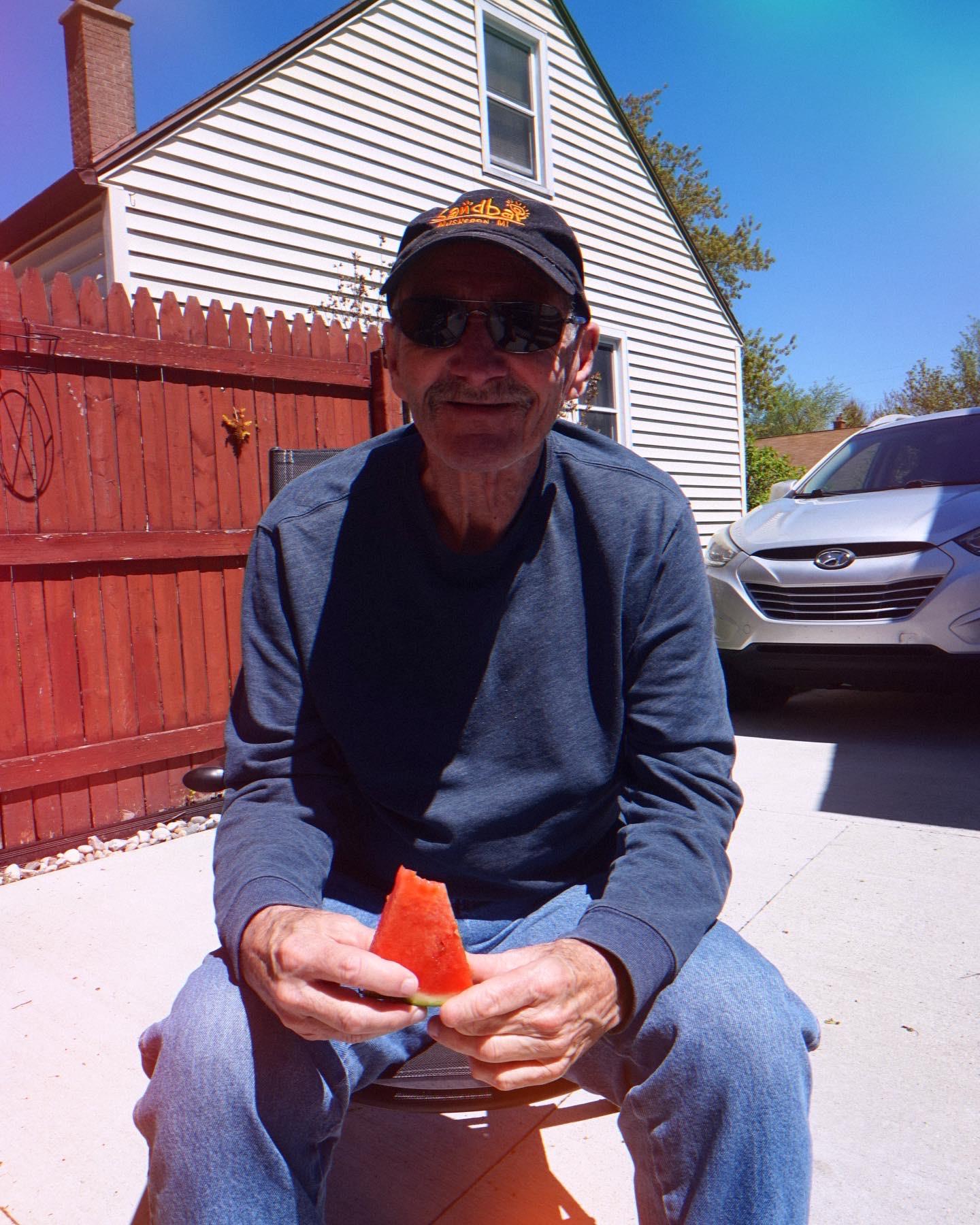A man sits in a yard holding a piece of watermelon, enjoying the warm weather in spring.