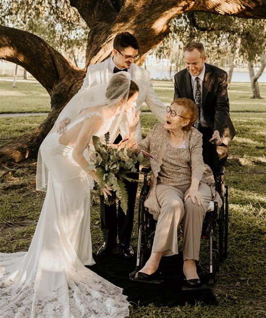 Bride in elegant gown warmly interacts with grandmother in wheelchair amidst family gathering.