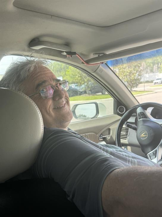 A man with glasses smiles warmly while driving his car on a bright day, relaxed and cheerful.
