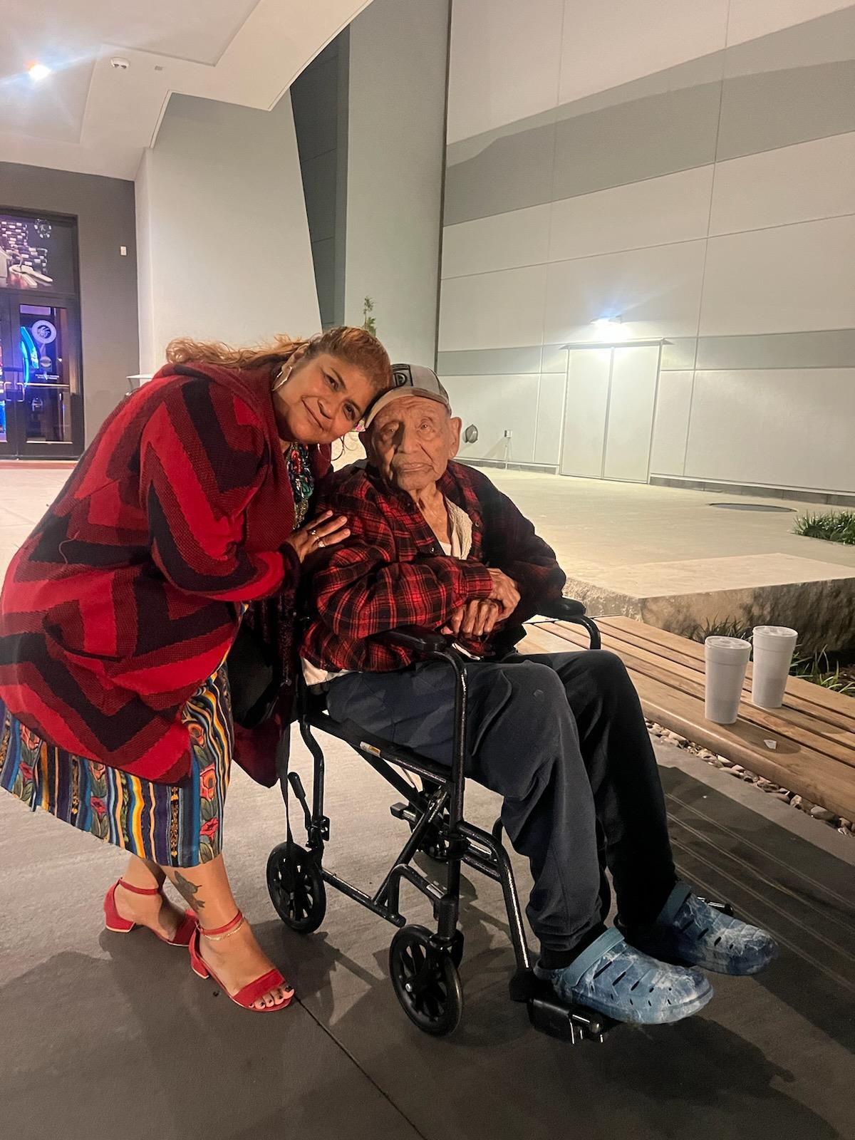 A woman smiles joyfully next to an elderly man in a wheelchair, enjoying the evening.