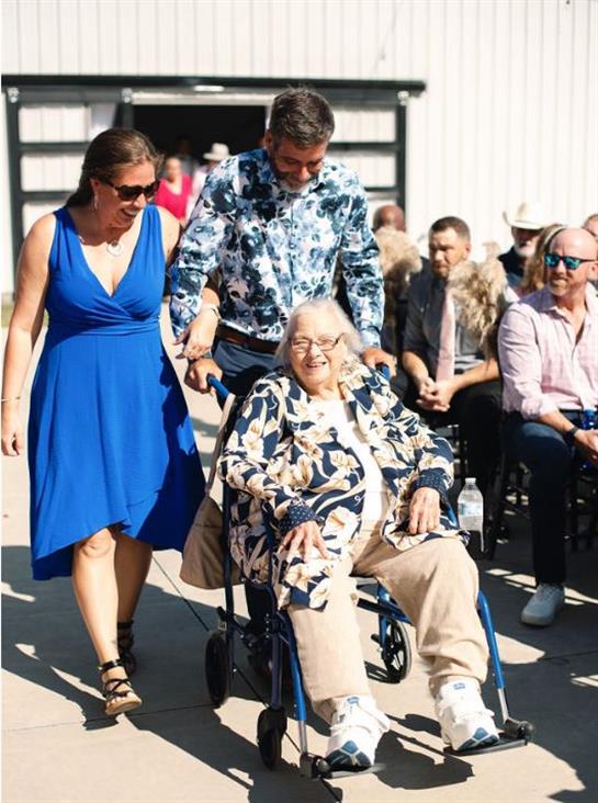 Two family members assist an elderly woman in wheelchair at a joyful outdoor gathering.
