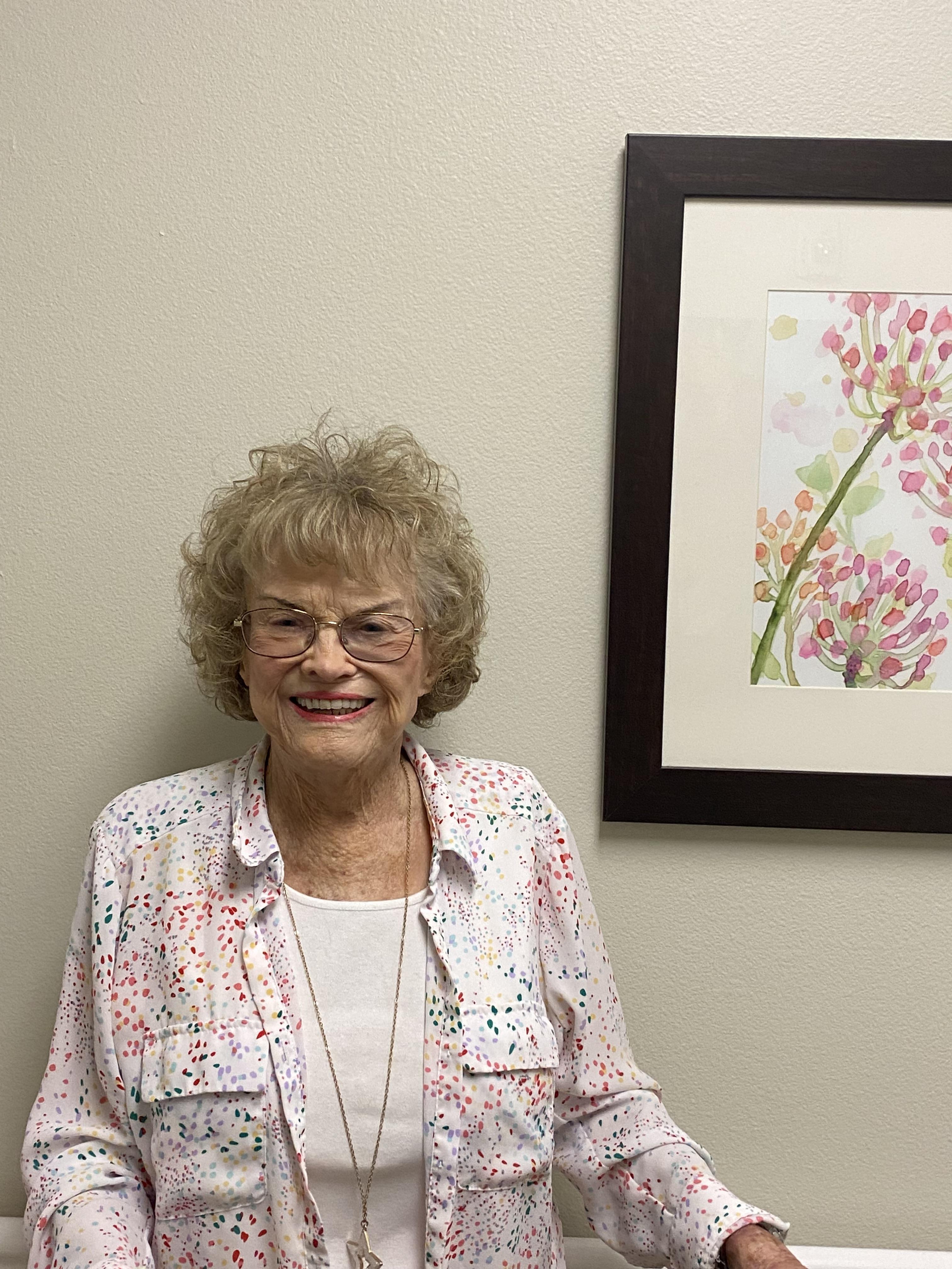 Elderly woman stands happily by a colorful floral painting in a well-lit interior.