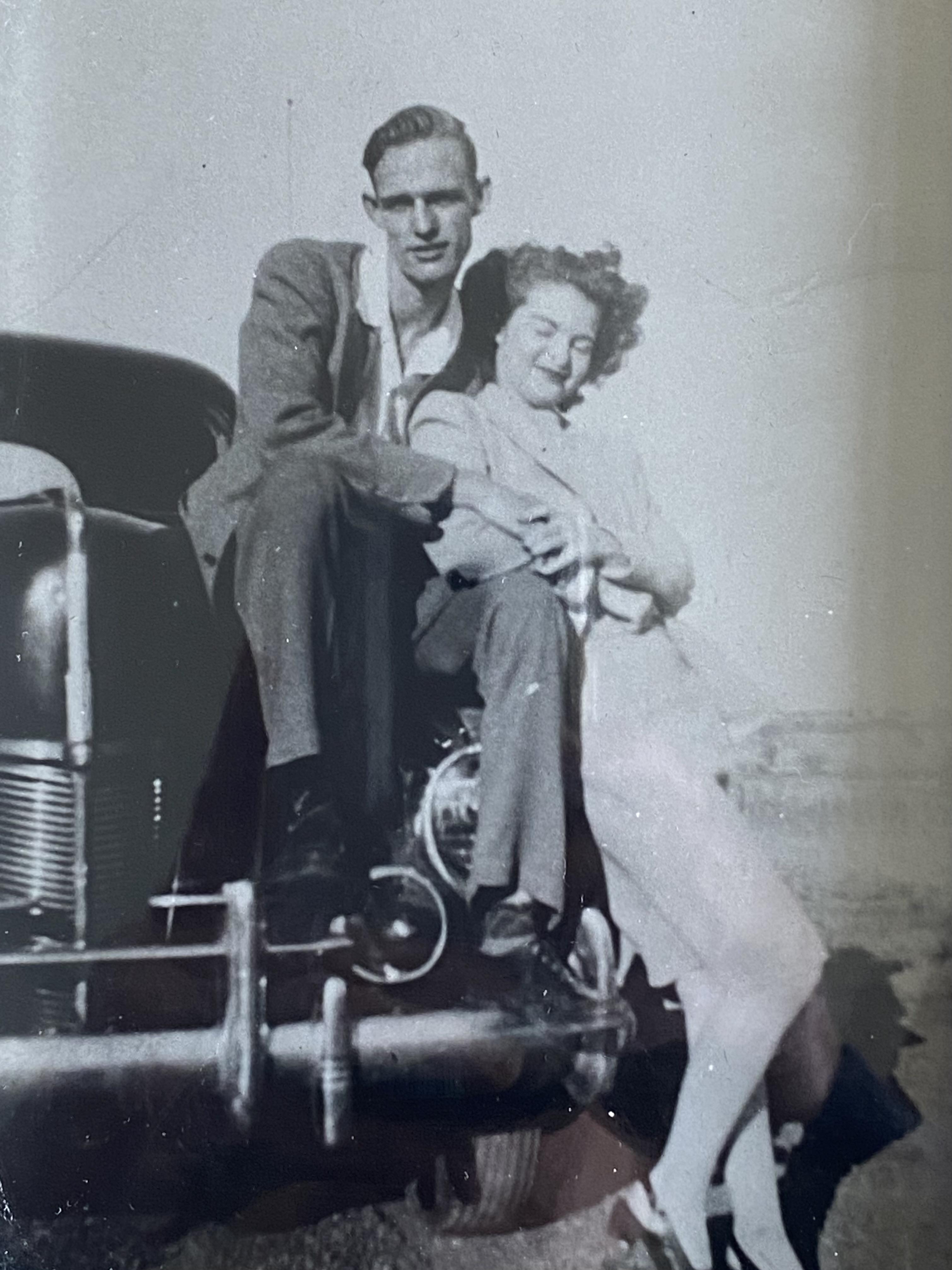 A man and woman pose happily on a classic automobile, enjoying a sunny afternoon.