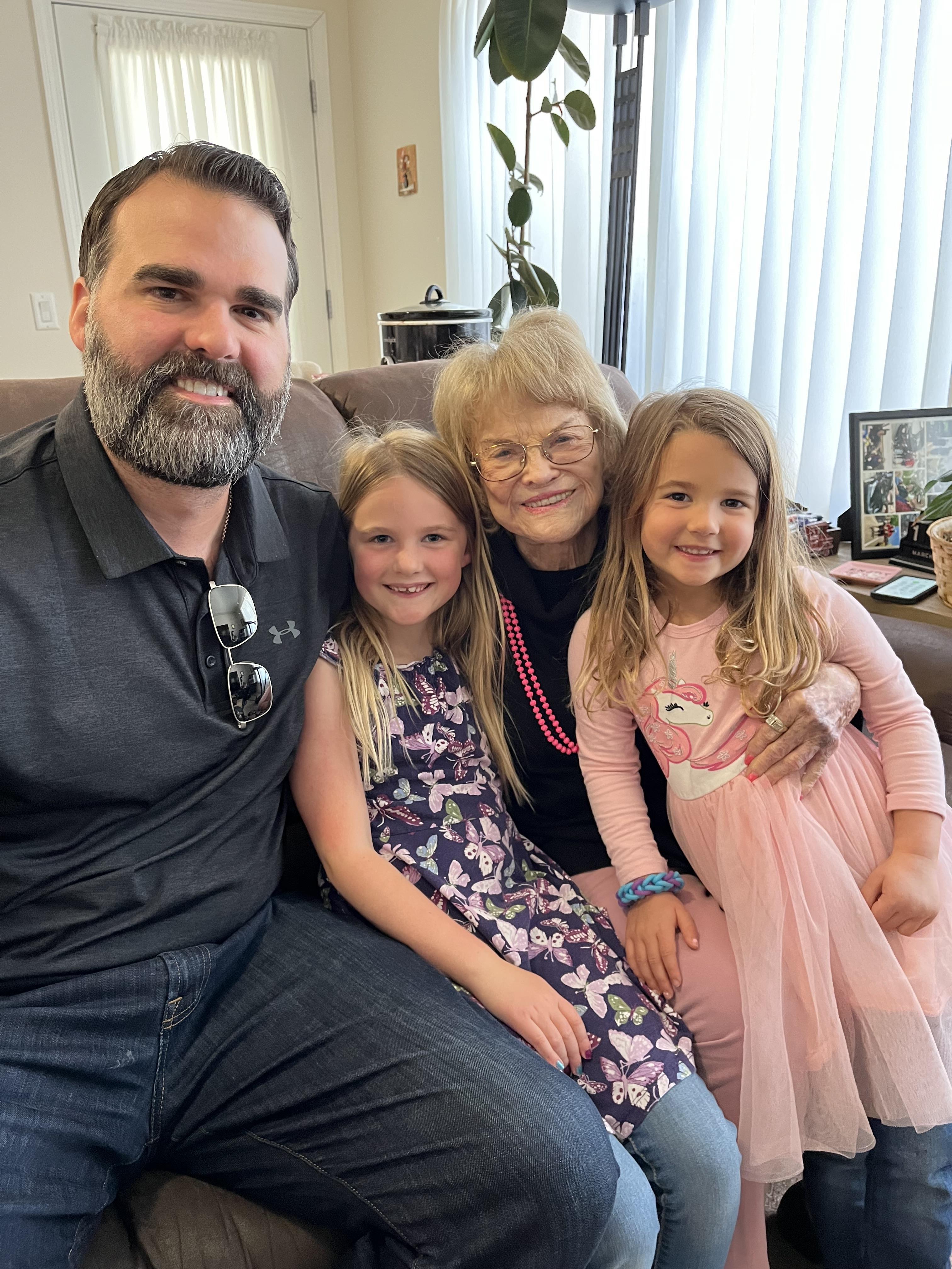 A man and two young girls sit beside a grandmother in a warmly decorated living room.