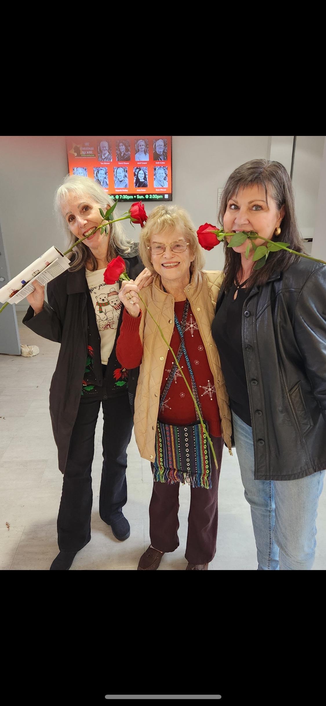Three women pose happily, each holding a rose, enjoying a joyful indoor celebration.