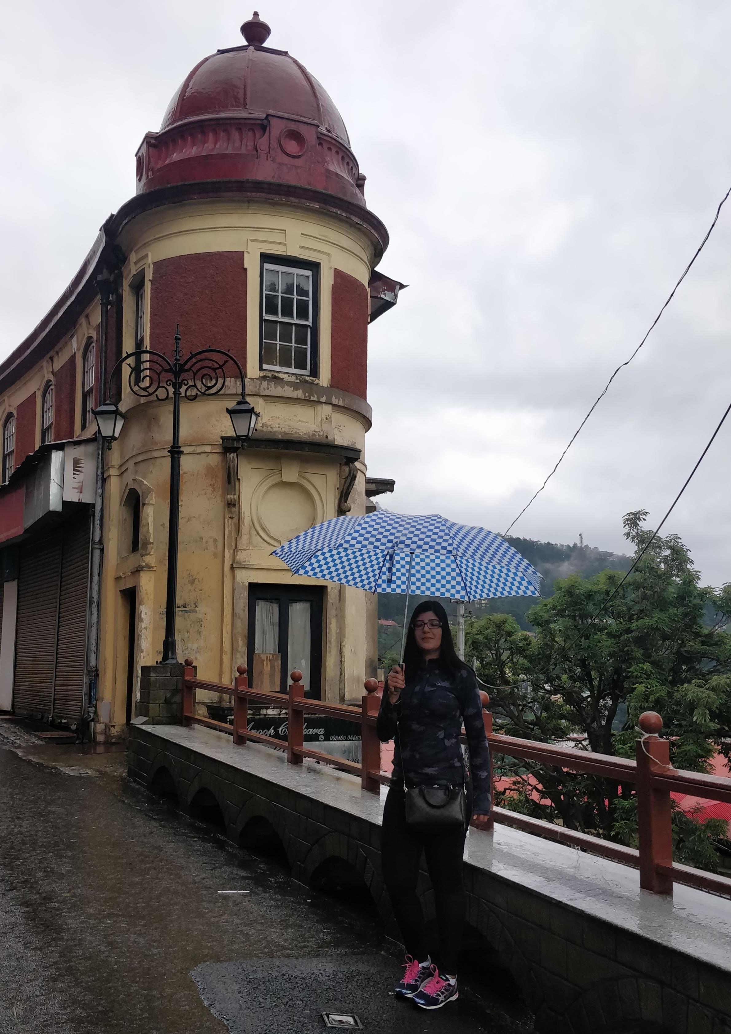 A woman stands by a colorful building, holding a blue umbrella while it rains.