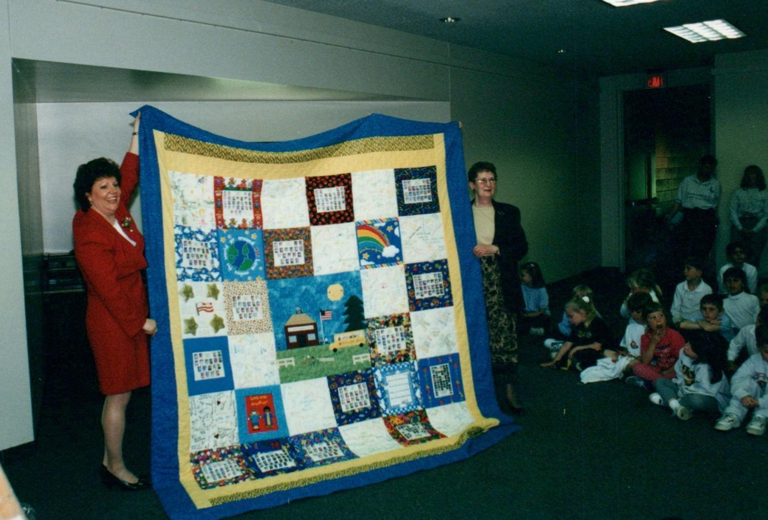 Two women showcase a handmade quilt to a captivated audience at a community gathering.