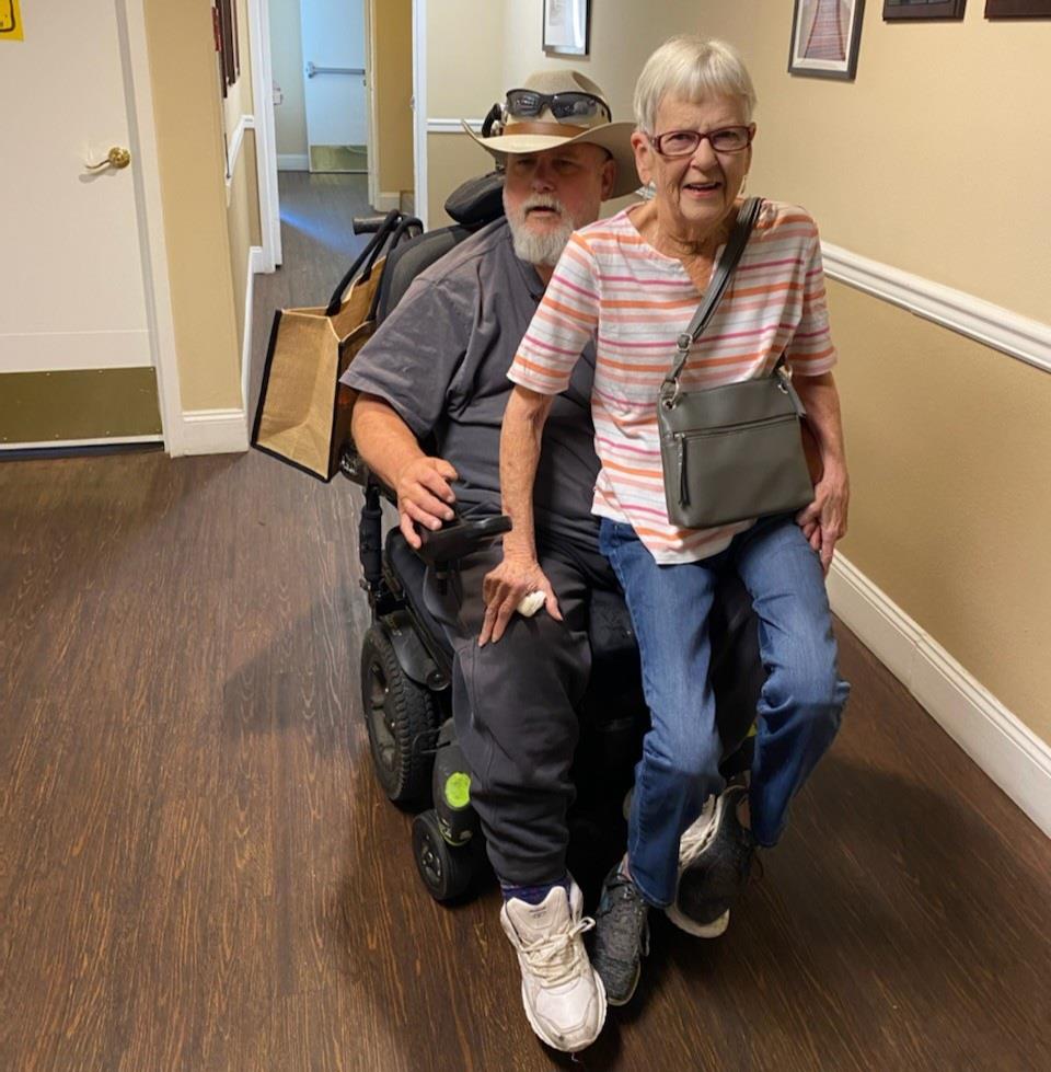 A man in a wheelchair rides with an elderly woman, both smiling in a care center hallway.