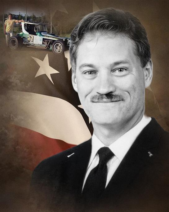 A mustached man smiles confidently with a racing car and American flag behind him.