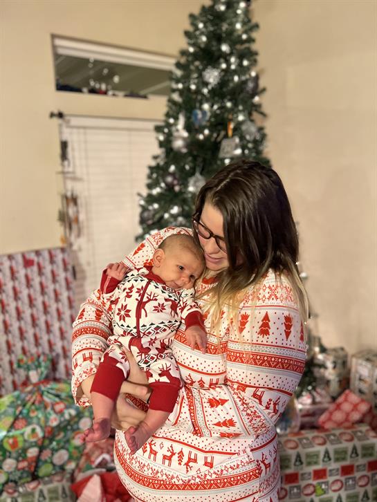 A woman holds her baby in festive attire, surrounded by gifts and a decorated Christmas tree.