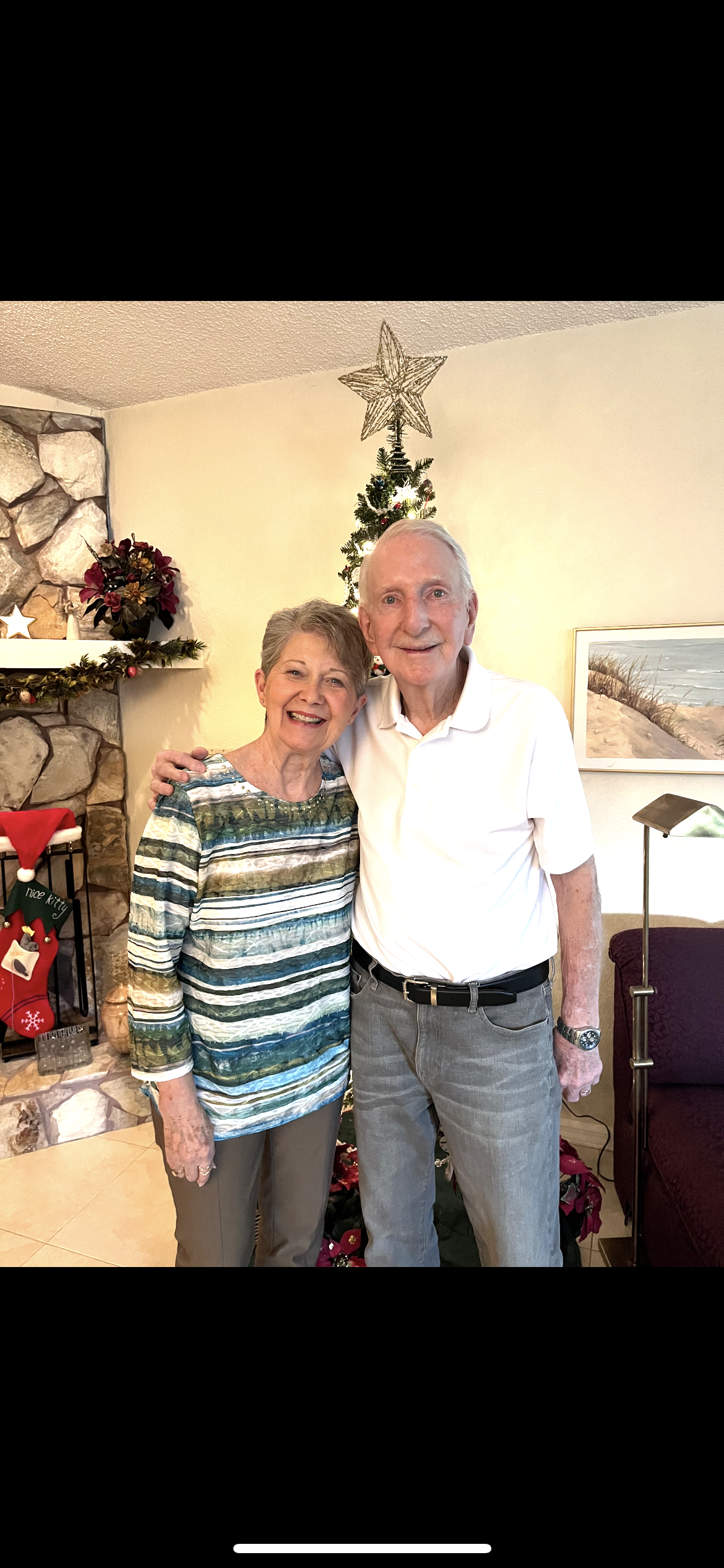 An elderly couple stands together, smiling in a warmly decorated living room for the holidays.