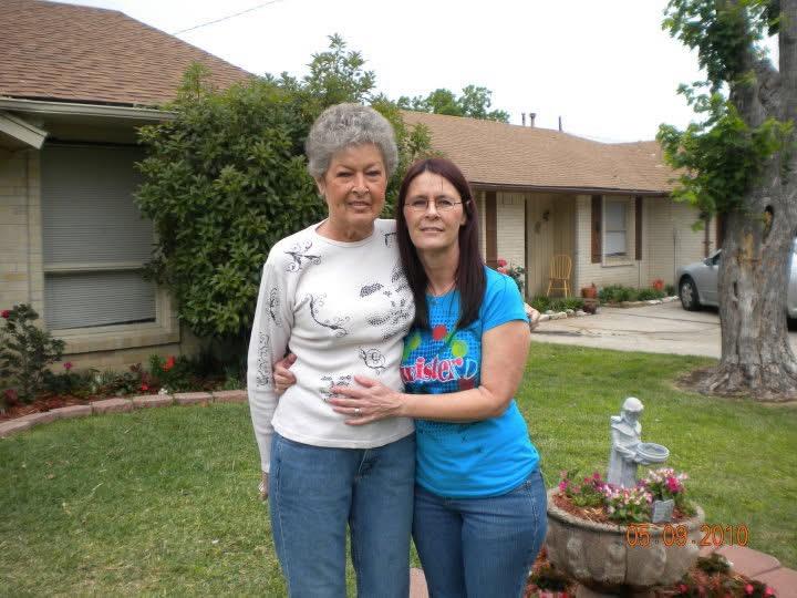 A granddaughter embraces her grandmother in a sunny garden outside their home, showcasing love.