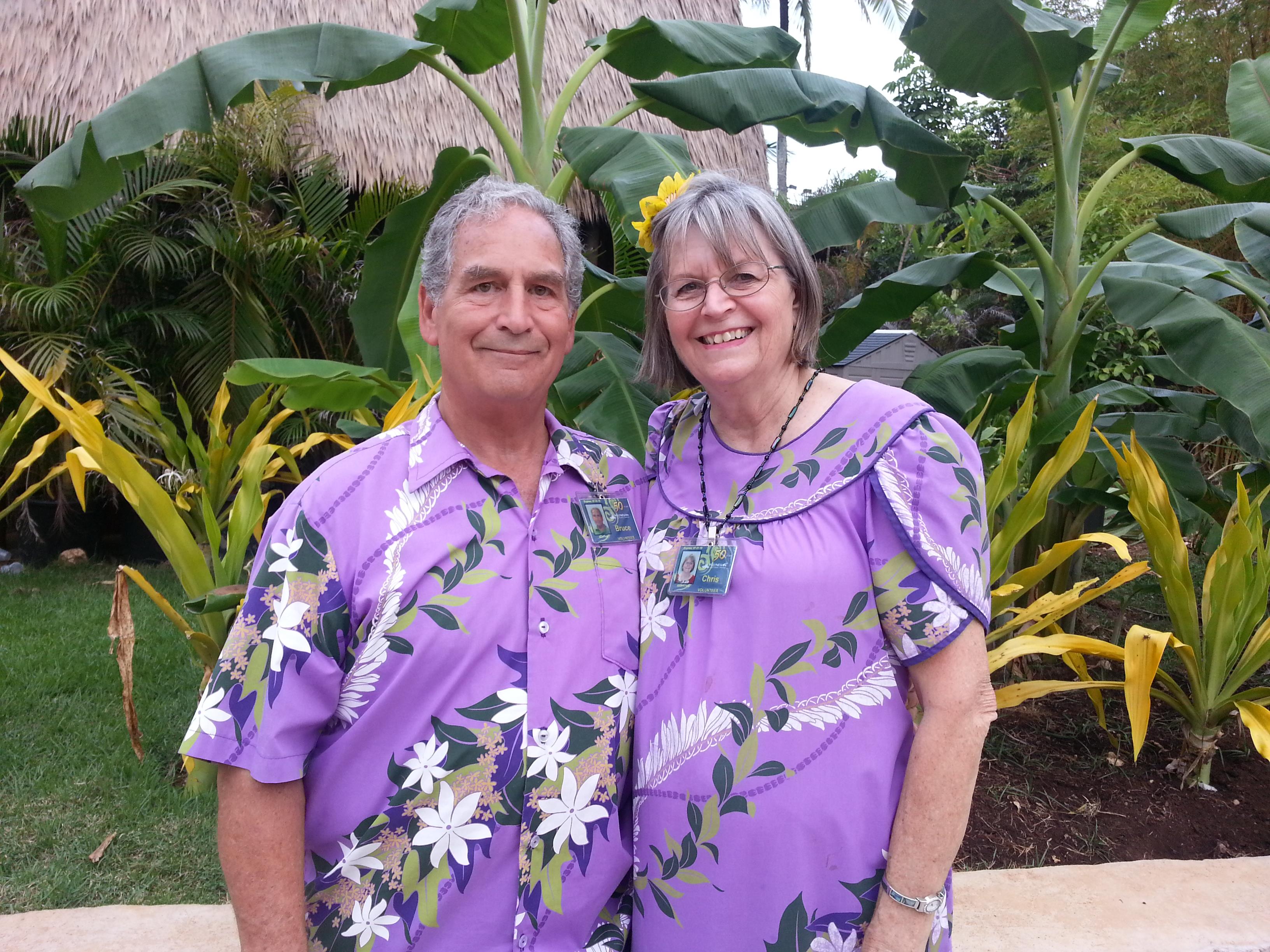A couple stands together in bright purple outfits surrounded by tropical plants under sunlight.