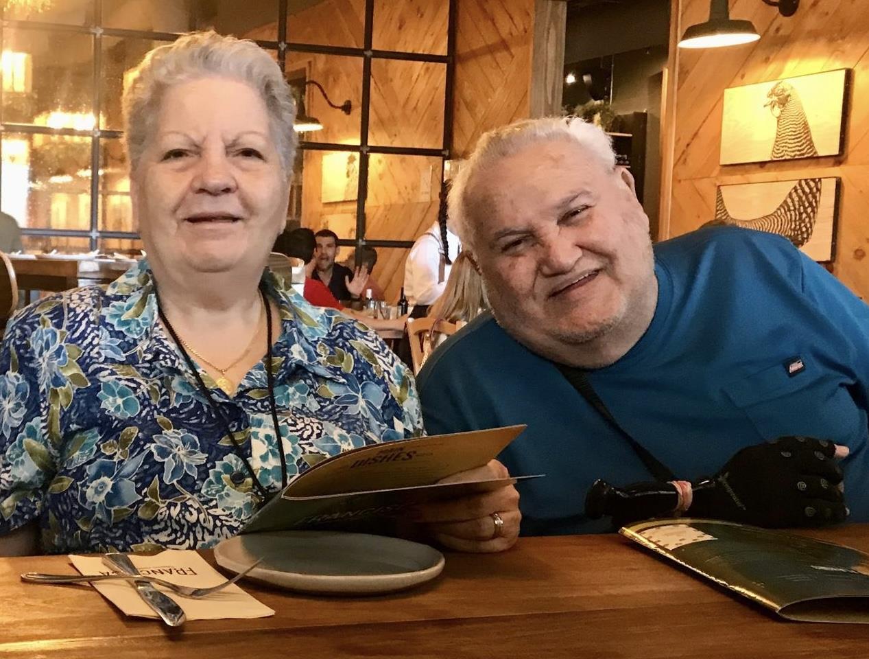 An elderly couple smiles while looking at menus in a busy eatery, surrounded by other patrons.