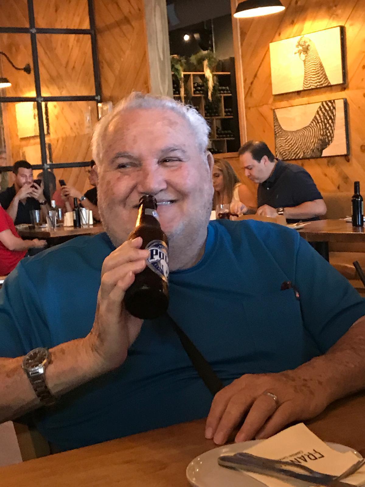 A man with white hair smiles while holding a bottle at a bustling restaurant with friends.