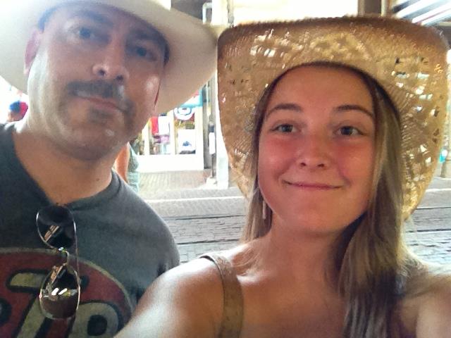 Two people take a selfie outside at a street fair, smiling and wearing hats under bright sunlight.