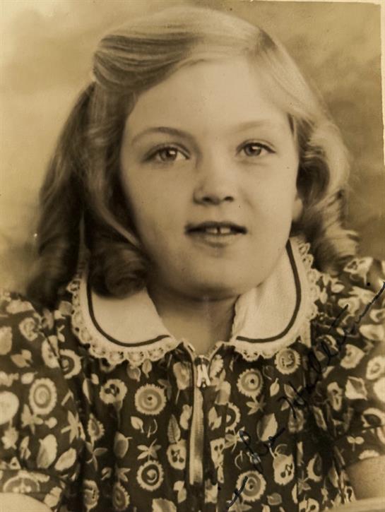 A young girl with curly hair wears a floral dress and smiles softly, embodying innocence and charm.