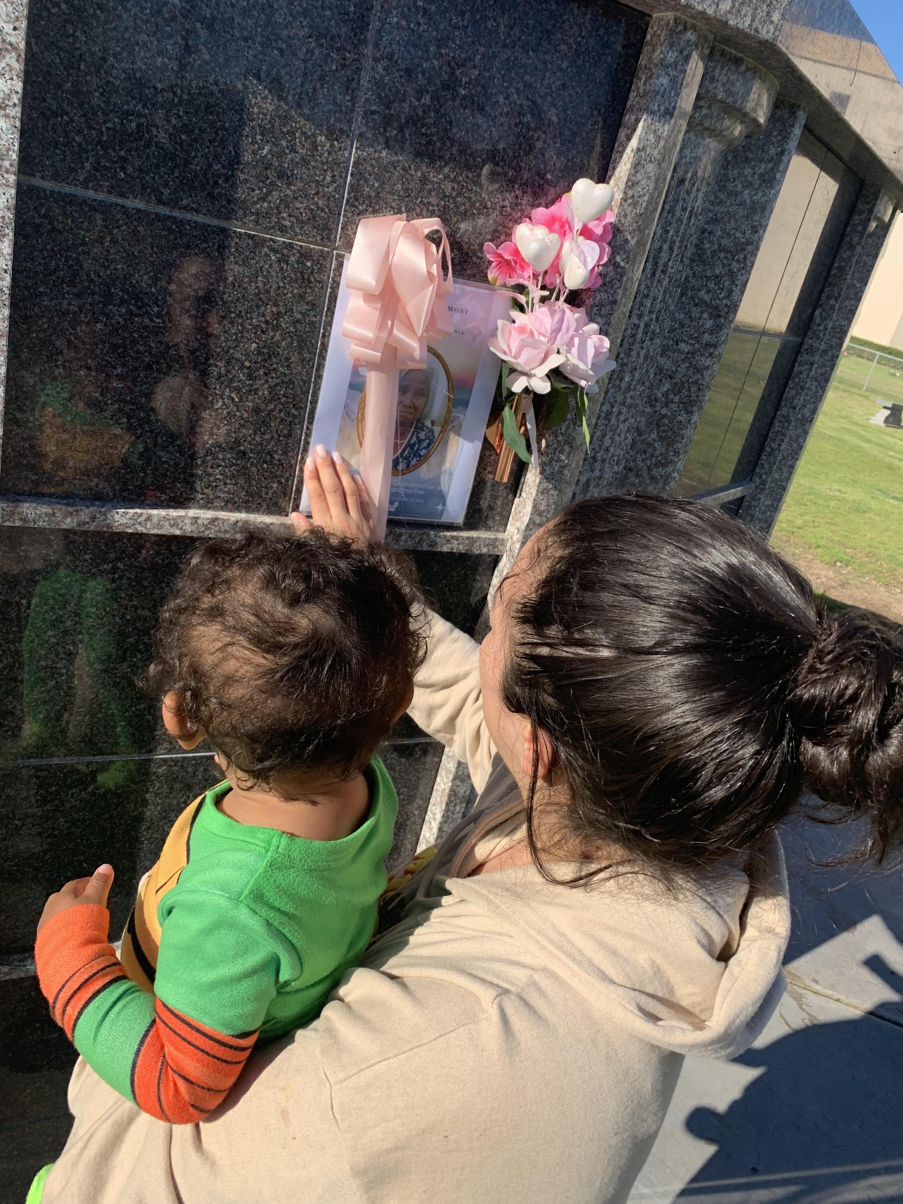 A woman holds a child while touching a memorial at a grave, surrounded by flowers, during daylight.
