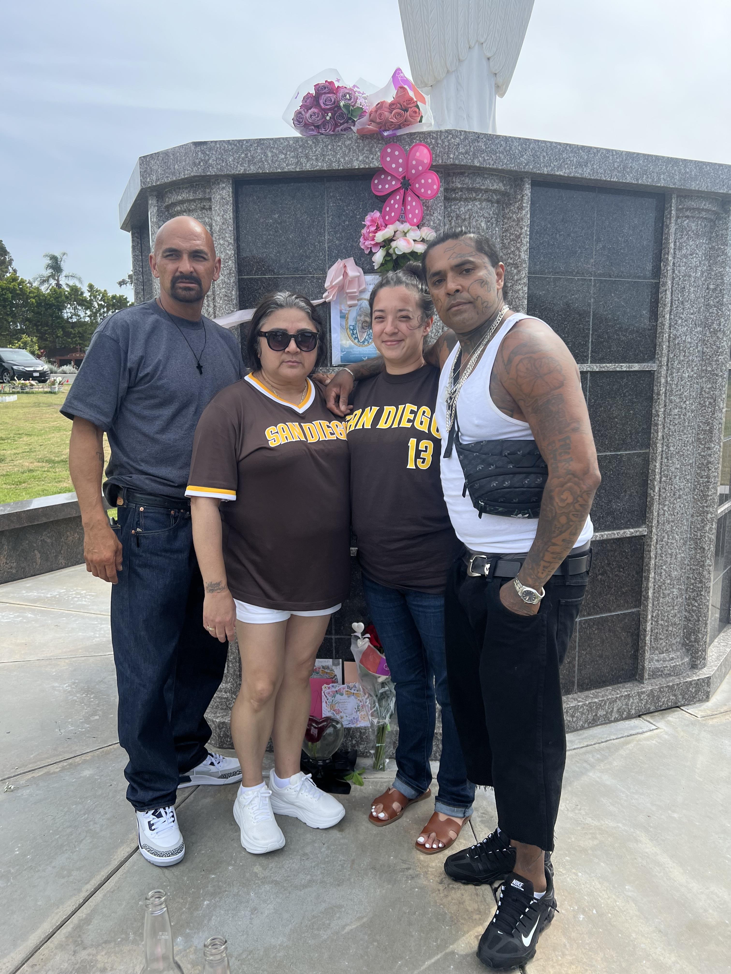 Group of four stands at a grave, paying tribute to a family member while holding flowers and drinks.