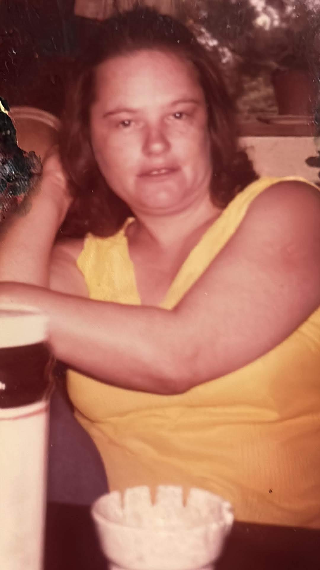 A woman with long hair is relaxing indoors, wearing a bright yellow top and smiling.