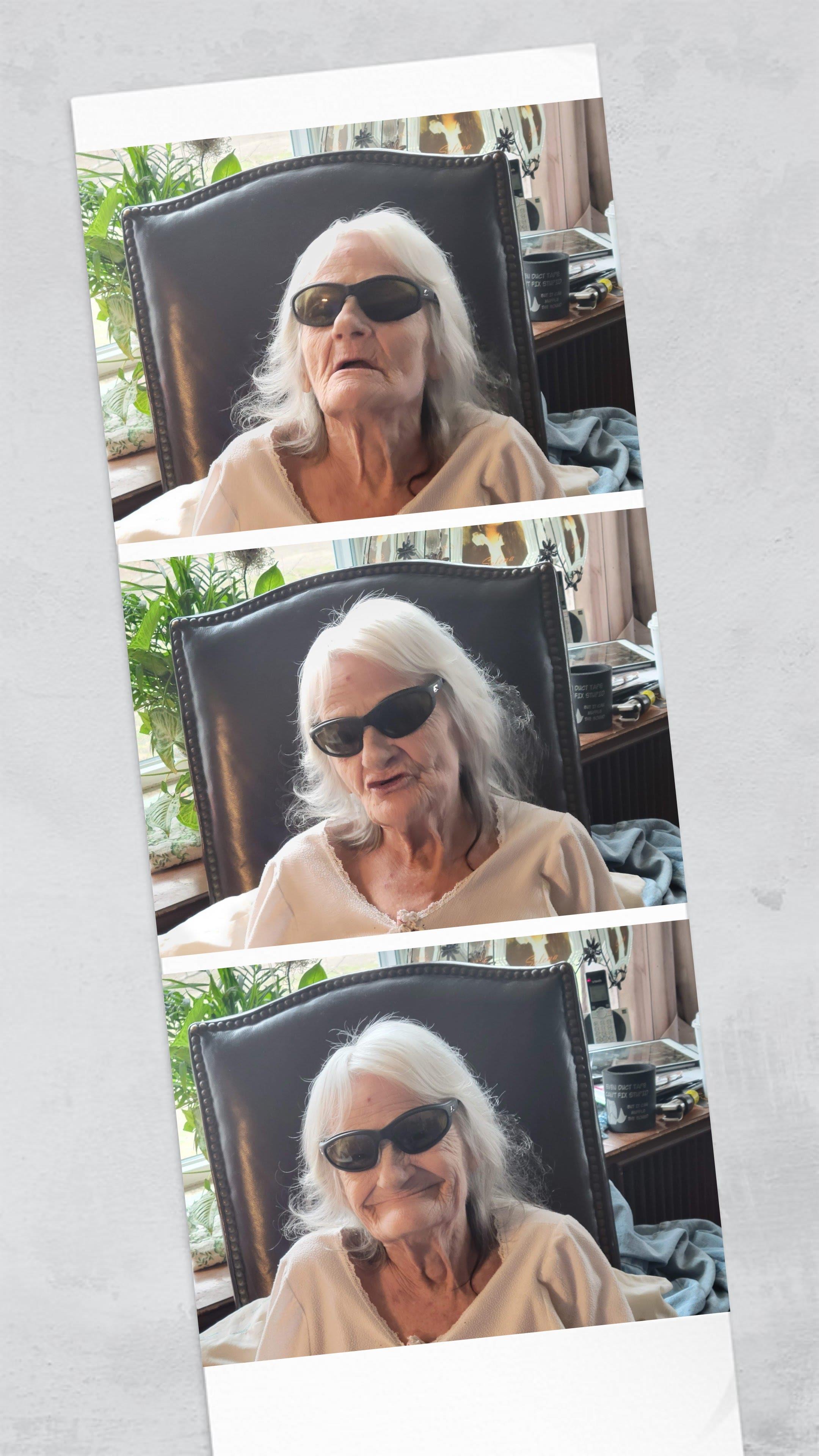 An elderly woman sits comfortably in a chair, wearing sunglasses and surrounded by greenery.