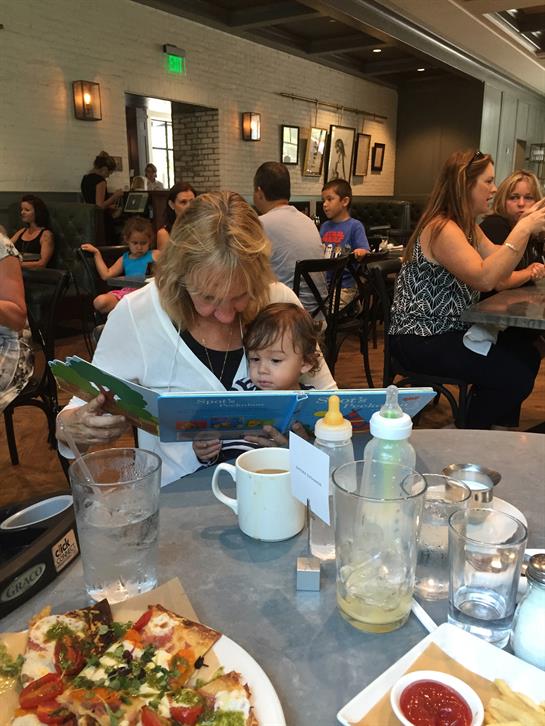 A woman reads a book to a young child at a bustling restaurant filled with families.