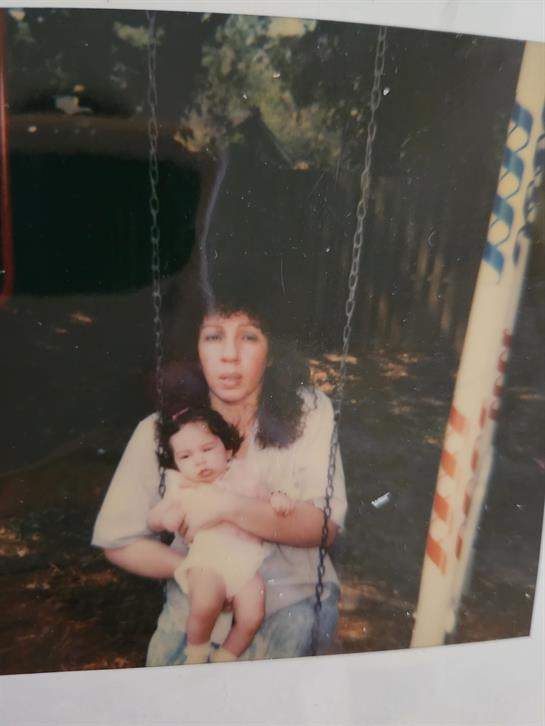 A mother holds her baby while seated on a swing in a park surrounded by trees.