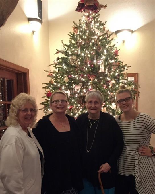 Four women stand together, smiling warmly in front of a festive Christmas tree.