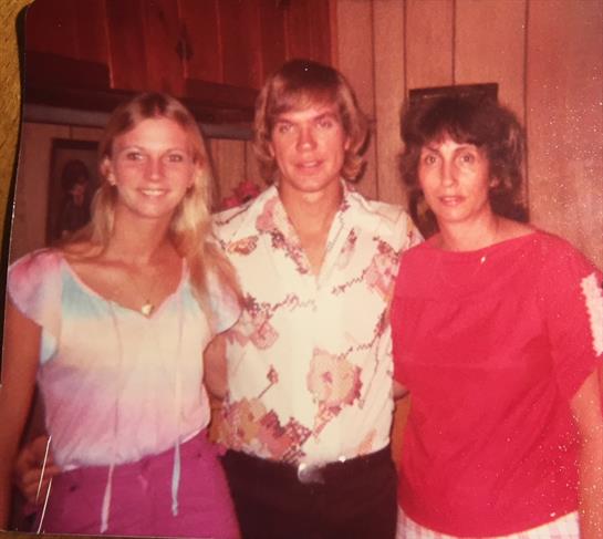 Three individuals smile while posing together in a cozy indoor setting from the 1970s.